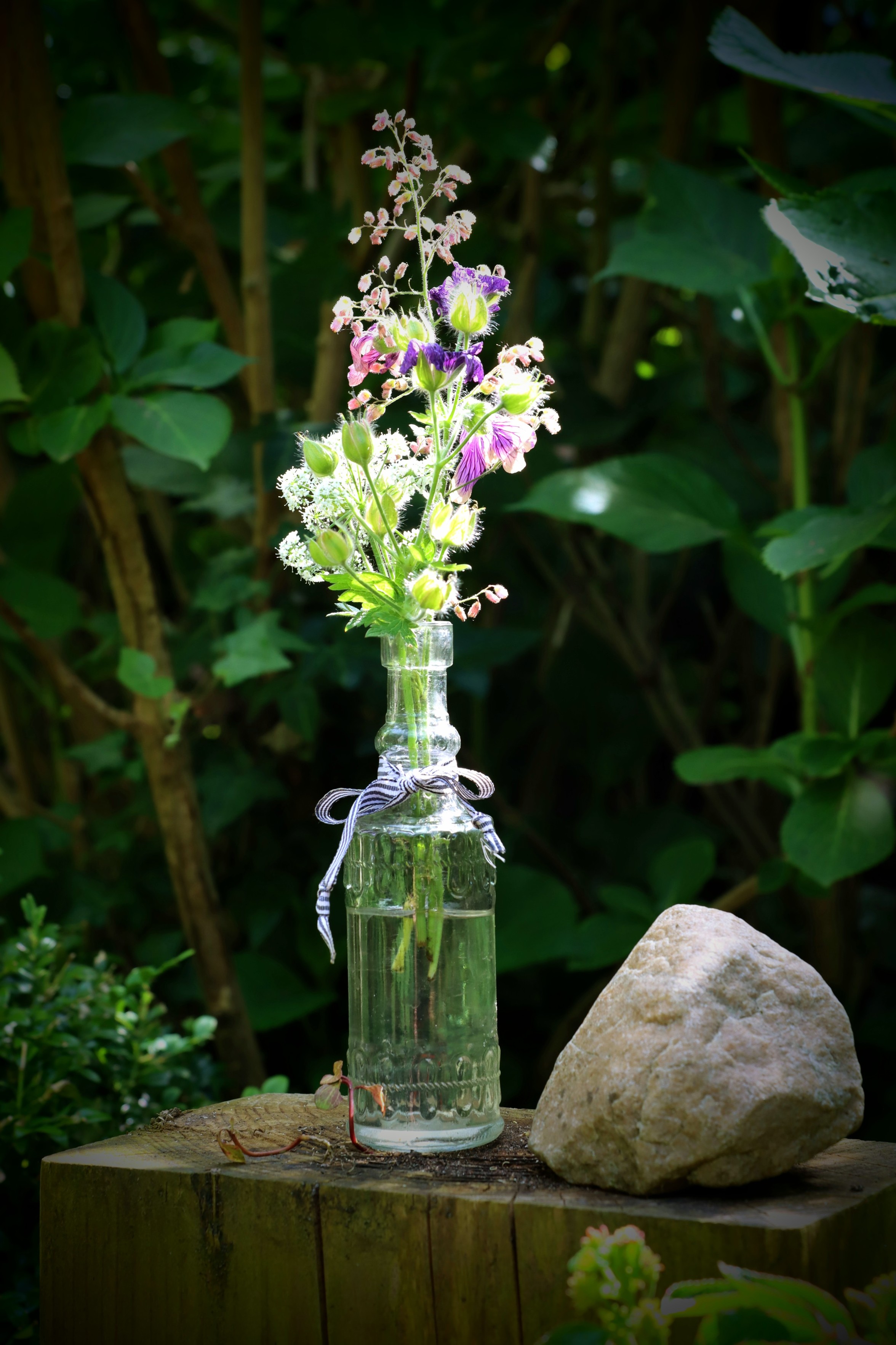Auf einem Holzklotz steht eine Glasflasche mit Schnittblumen. Um den Flaschenhals ist eine Schleife gebunden. Neben der Flasche, auf dem Holz liegt ein Stein. Auf einem Holzklotz steht eine Glasflasche mit Schnittblumen. Um den Flaschenhals ist eine Schleife gebunden. Neben der Flasche, auf dem Holz liegt ein Stein.