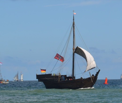 Kogge: Einmastiges Segelschiff, hölzerner Rumpf, schräger Bug, erhöhter Heckaufbau, segelt unter gerefftem Rahsegel dicht unter Land.