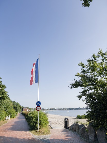 Zugang zum Strand, Strand und Ostsee, Weg an der Promenade, Flaggenmast mit Schleswig-Holstein-Flagge, blauer wolkenloser Himmel. 