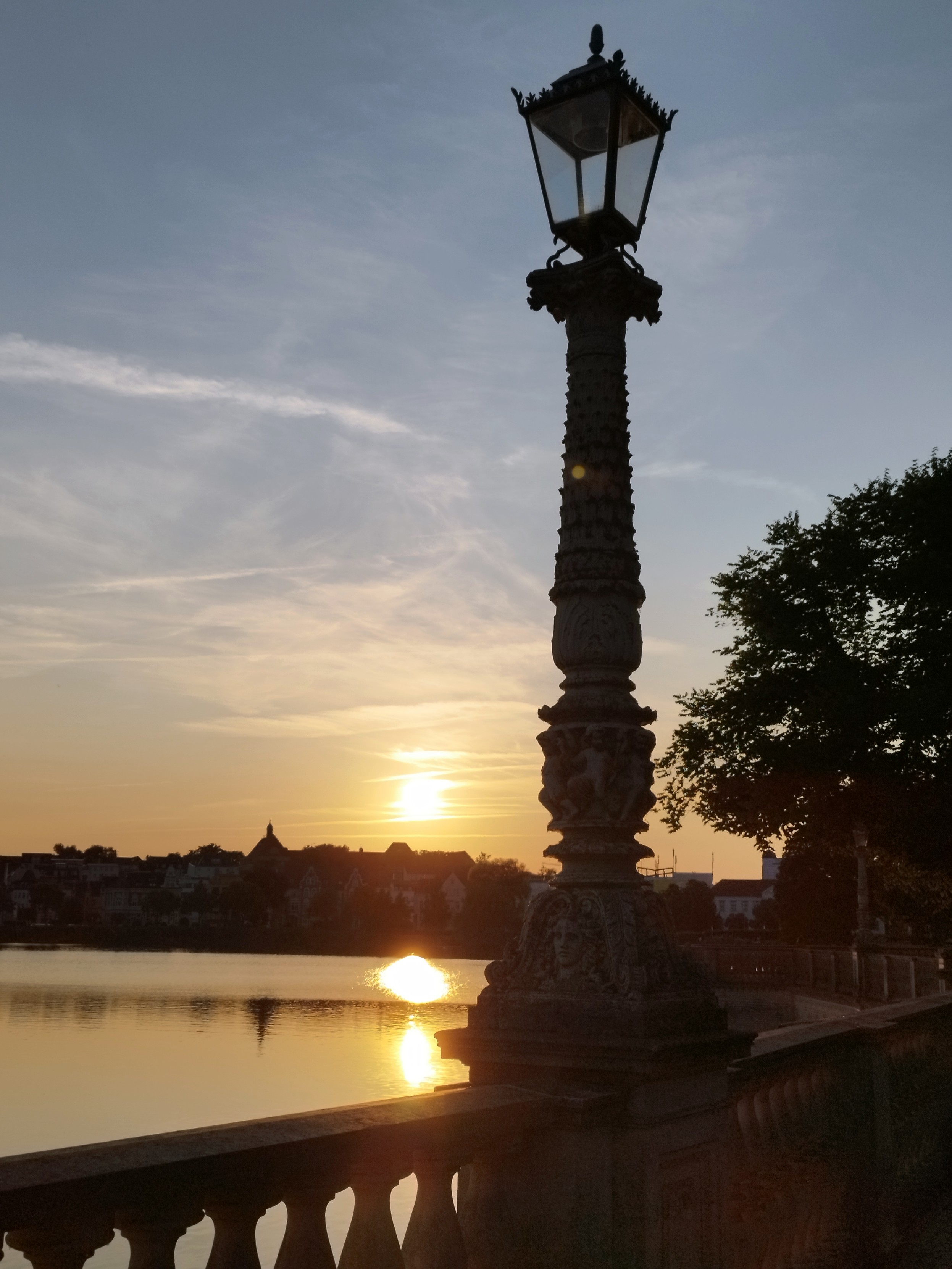 Ein Sonnenuntergang über einer Stadtsilhouette an einem See. Im Vordergrund eine Laterne an einem Brückengeländer. Rechts steht ein Baum.