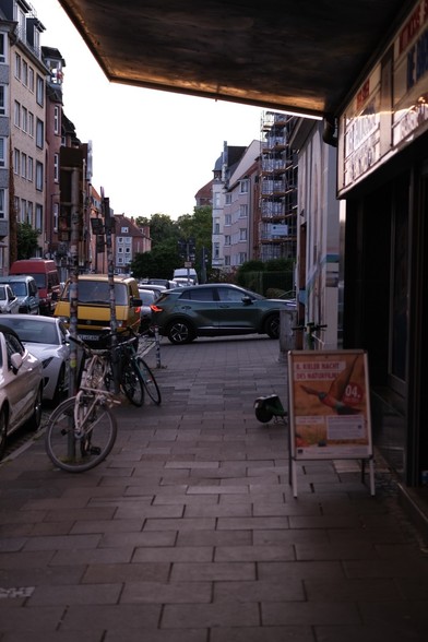 Das Foto zeigt eine ruhige Straßenszene in einer Stadt neben dem Studio Kino in Kiel. Im Vordergrund ist ein Gehweg aus Steinplatten zu sehen, auf dem zwei Fahrräder an Straßenschlidern lehnen. Ein Schild mit der Aufschrift "8. KIELER NACHT DES NATÜRFILMS" steht am rechten Rand des Bildes. Dahinter steht ein Leihroller. Im Hintergrund sind mehrere Gebäude zu sehen, darunter Wohnhäuser mit Balkonen und ein Gebäude mit Gerüst. Parkplätze entlang der Straße sind mit verschiedenen Autos und einem B…