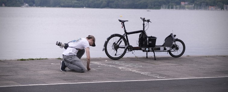 Eine Person kniet auf einem Weg am Wasser und schreibt mit Kreide auf den Gehweg. In der Nähe befindet sich ein Fahrrad mit Ladefläche. Der Hintergrund zeichnet sich durch ein ruhiges Gewässer und Grün in der Ferne am Kieler Ostufer aus.