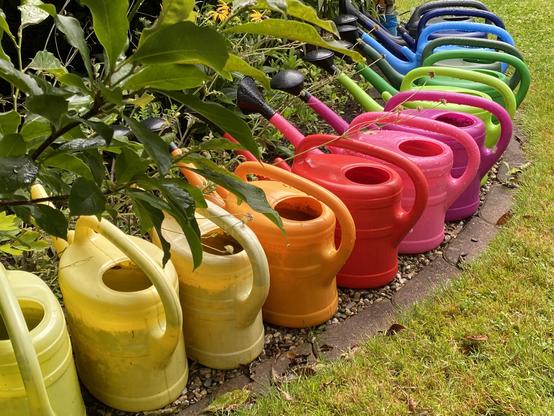 13 Gartengießkannen in Regenbogenfarben, im Halbkreis an einer Rasenkante aufgereiht.