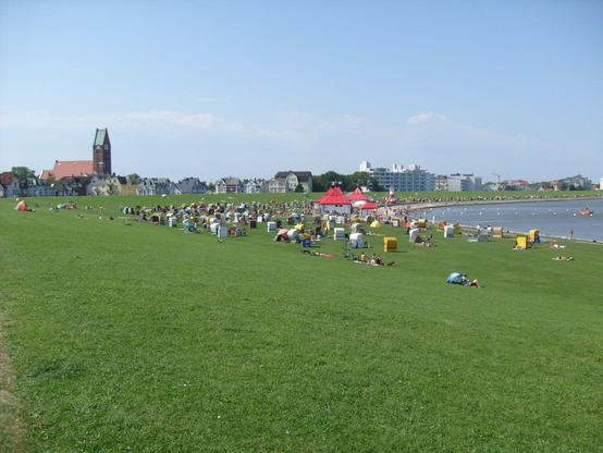 Grünstrand mit Strandkörben. Im Hintergrund links eine Kirche, rechts Hotels.