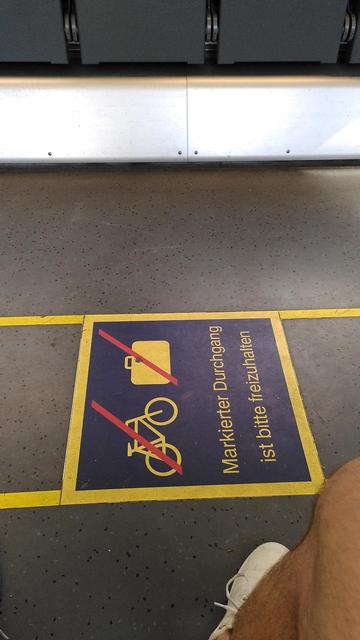 Auf dem Boden im Fahrradbereich eines RE70 sind Markierungen um die Abstellbereiche links und rechts des Ganges angelegt. In ihnen sind ein durchgestrichenes Fahrrad und ein durchgestrichener Koffer abgebildet. Dazu der Text "Markierter Durchgang ist bitte freizuhalten"
