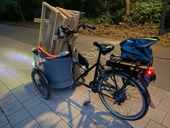 im Dämmerlicht: ein Lasten Dreirad, das in der Wanne vorn sechs senkrechte Holzrahmen mit Plexiglasfüllung hat.