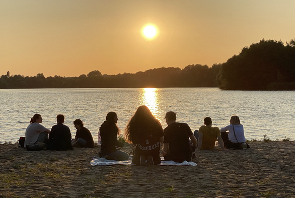 Sonnenuntergang über dem Eichbaumsee. Im Vordergrund die Silhouetten von Menschen, die am Strand sitzen. Das Foto ist dunkler als die Wirklichkeit. Es wirkt sehr ruhig und chillig. Die laute Musik hört man ja nicht…