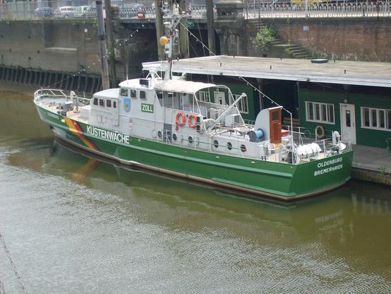 Zollkreuzer an einem Ponton festgemacht. Großes Boot bzw. kleines Schiff. Grüner Rumpf, Aufschrift "Küstenwache", graue Aufbauten. 