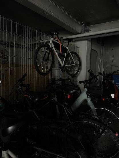 In einer vollen Fahrradgarage hängt ein Gravel mit Lenker und Pedale an einem Zaun über den anderen Rädern.