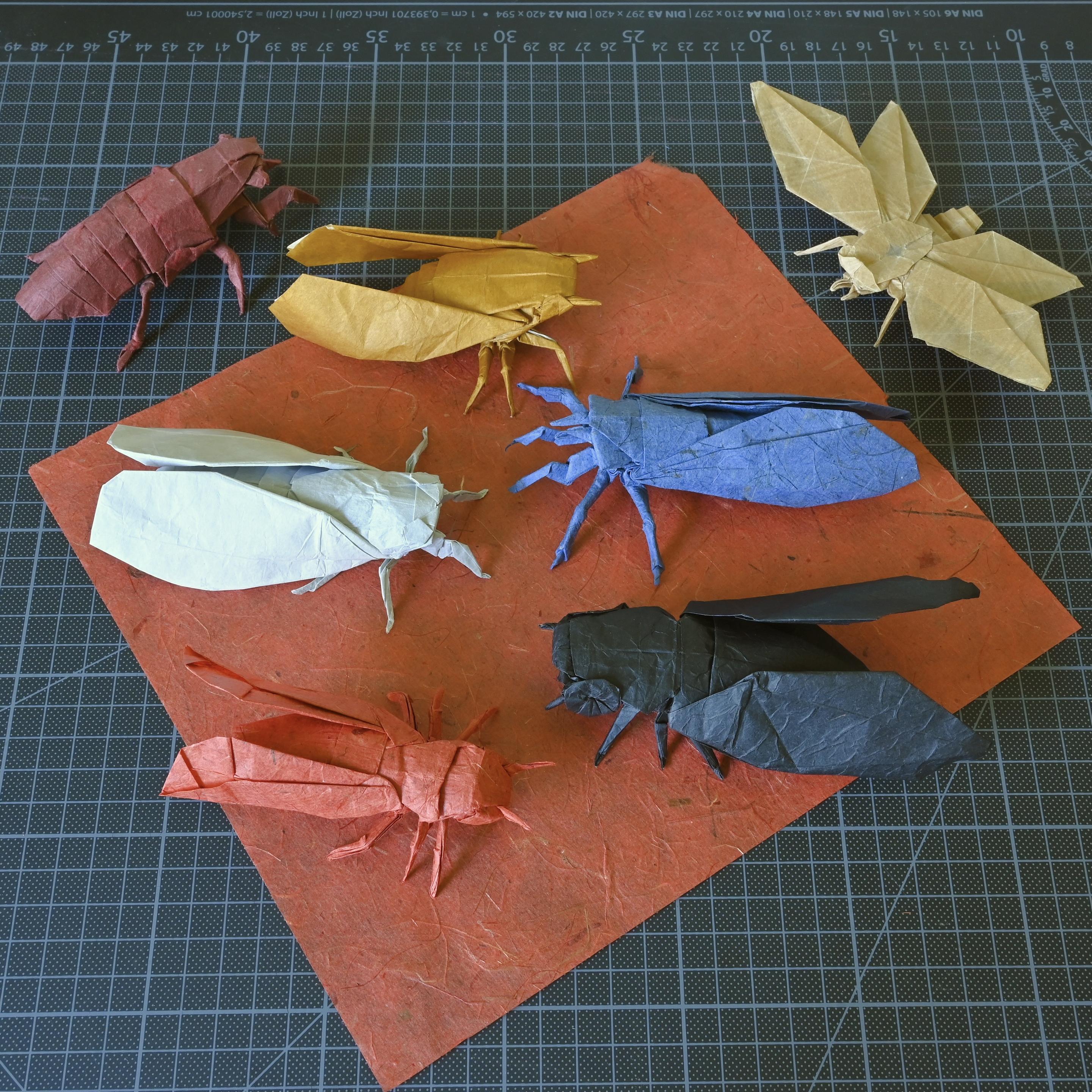 Seven colorful origami cicadas, arranged on a red paper square over a black cutting mat.