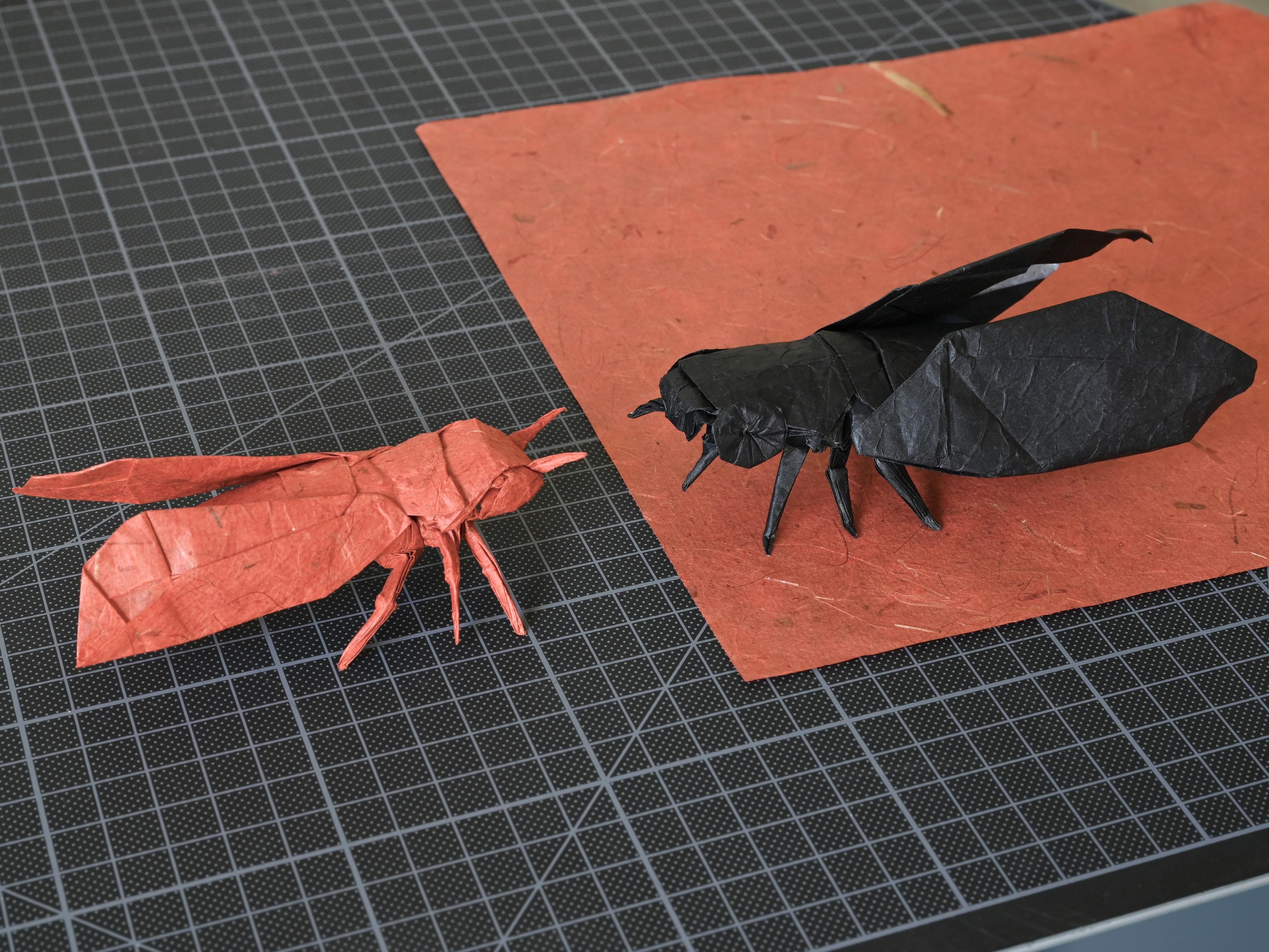 Two origami cicadas, one red and one black, displayed on a black cutting mat with a piece of red paper beneath the black model.