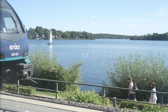 Blick aus einem Zug vorbei an einem anderen Zug auf einen See, vor den zwei Wanderer*innen vorbei spazieren und auf dem ein Segelboot unterwegs ist.