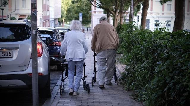 Ein paar mit Rollatoren spaziert nebeneinander auf einem Fußweg. Links von ihnen ist eine Reihe Parkplätze, rechts von ihnen ist eine Hecke (die zu einem großen Parkplatz gehört)