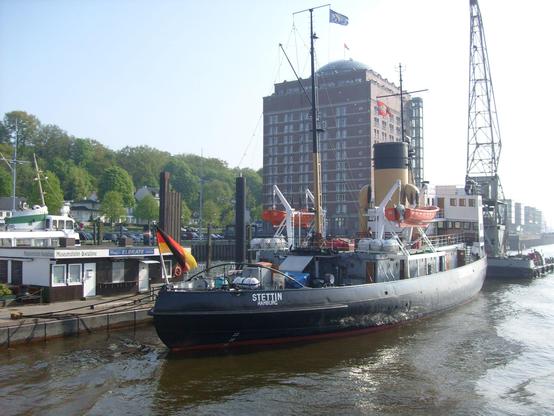 Eisbrecher STETTIN im Museumshafen Övelgönne in Hamburg. Ein Schiff mit schwarzem Rumpf und hohem gelben Schornstein liegt an einem Ponton. Im Hintergrund ein Hochhaus mit Backsteinfassade und einer Glaskuppel.