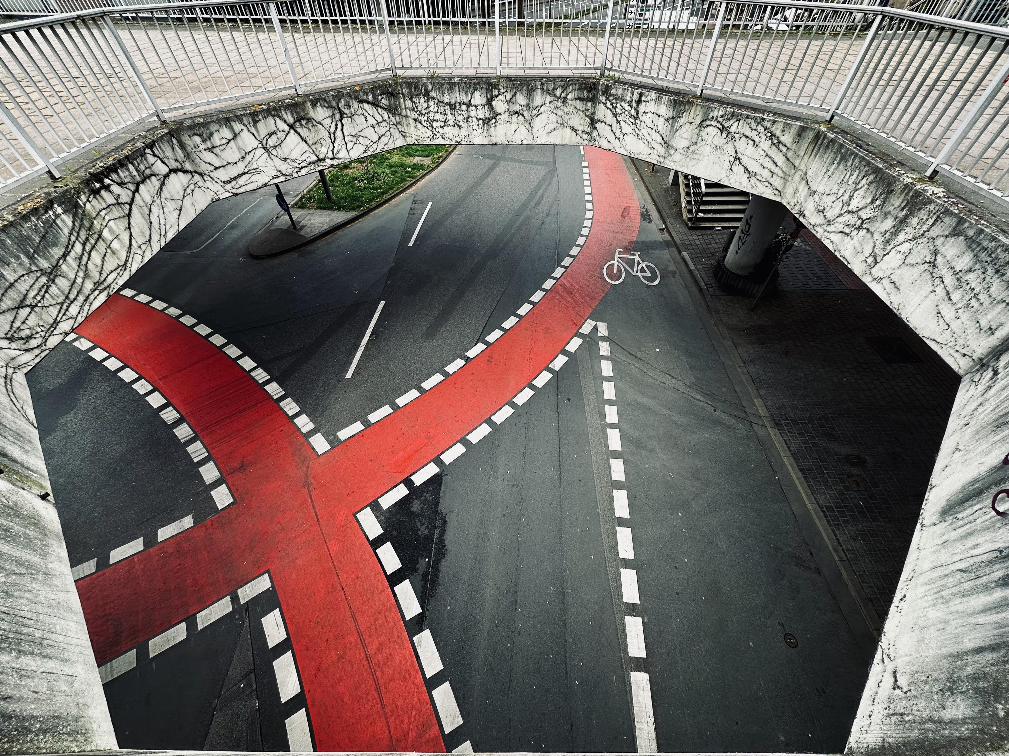 A road intersection viewed from above, featuring a distinctive red X-shaped bike lane. The bike lane is bordered by dashed white lines. A white bicycle symbol is visible on the pavement, indicating a cyclist route. The intersection is surrounded by a concrete bridge with white railings, providing an elevated perspective. The area has visible markings and weathered textures on the concrete surfaces. A road intersection viewed from above, featuring a distinctive red X-shaped bike lane. The bike lane is bordered by dashed white lines. A white bicycle symbol is visible on the pavement, indicating a cyclist route. The intersection is surrounded by a concrete bridge with white railings, providing an elevated perspective. The area has visible markings and weathered textures on the concrete surfaces.