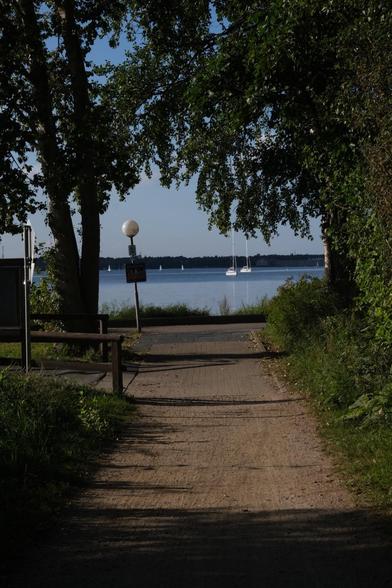 Ein Weg, der durch Bäume zu einem ruhigen Gewässer führt. Zwei Segelboote sind auf dem Wasser zu sehen, mit einem klaren blauen Himmel darüber.
