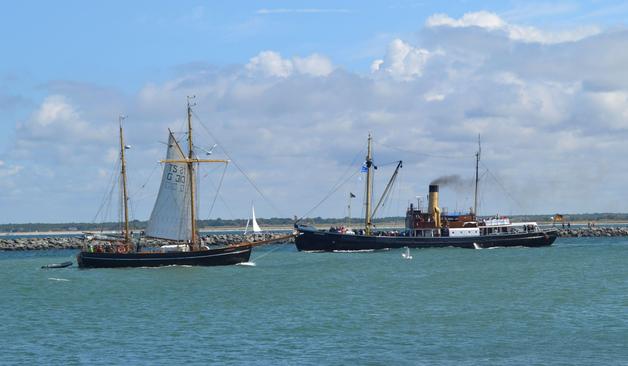 Ein von linkskommendes Segelschiff mit schwarzen Rumpf, das am vorderen Mast Segel gesetzt hat, begegnet einem von rechts kommenden Dampfschiff mit schwarzem Rumpf und rauchenden Schornstein. Im Hintergrund eine Mole. 