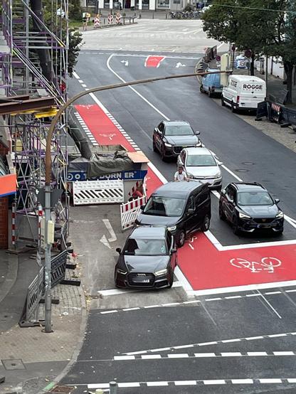 Auf einer neuen Kreuzung mit Baustellenanteilen steht ein Auto quer über den Radweg in Mittellage und blockiert diesen. Der Rechtsabbieger auf den der Autofahrer fahren wollte ist bereits vollständig belegt und so steht das Auto auf dem Radweg und teilweise auch auf dem Streifen für den Geradeausverkehr. Hinter dem Auto steht ein Radfahrer und kommt nicht vorbei.