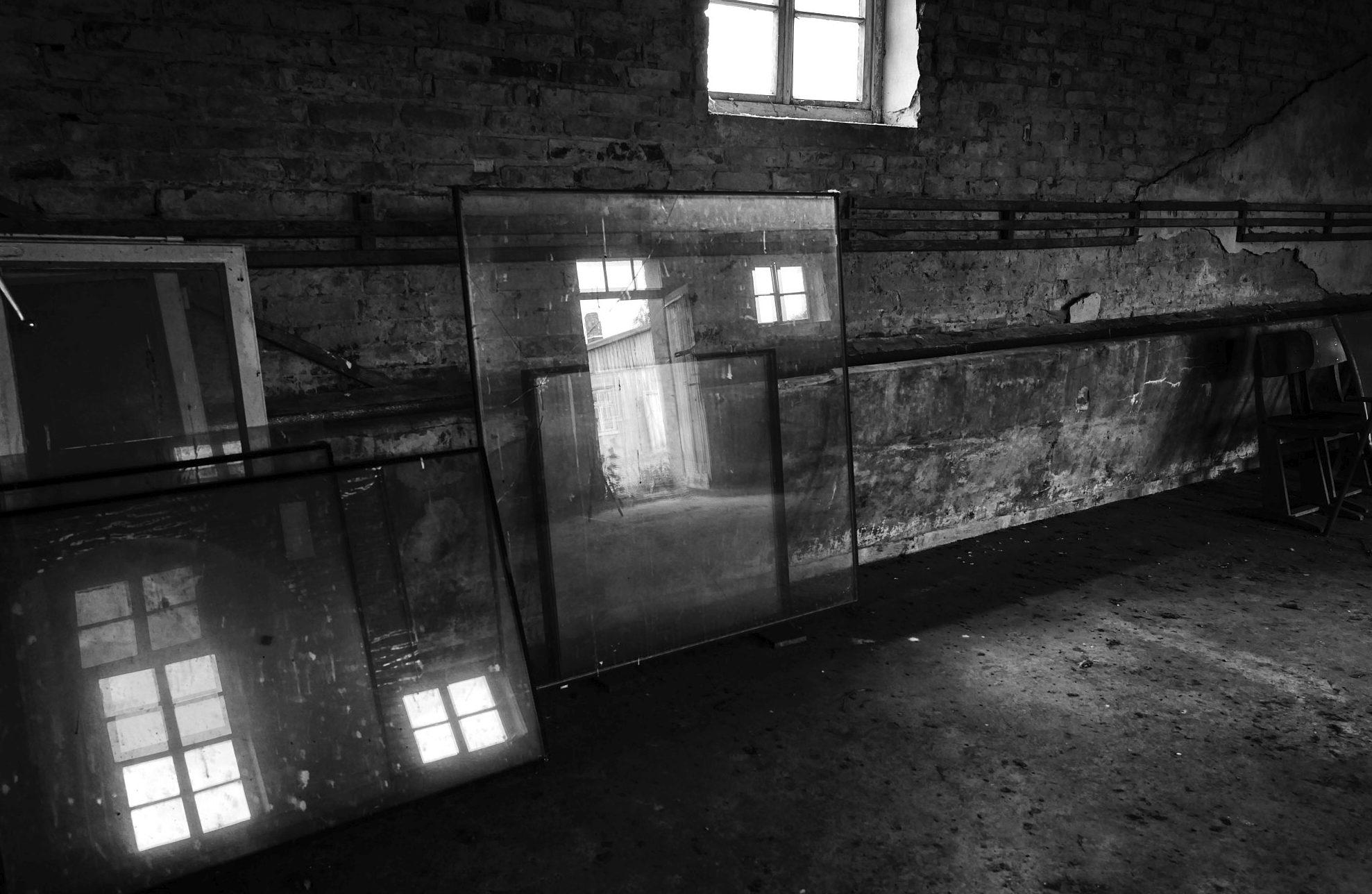 B&W-Bild mit der Innenansicht eines ehemaligen Waschraums. In dem Raum lagern einige Glasscheiben und warten auf ihre Verwendung. In ihnen spiegeln sich diverse Fenster und ein Eingangstür. 


