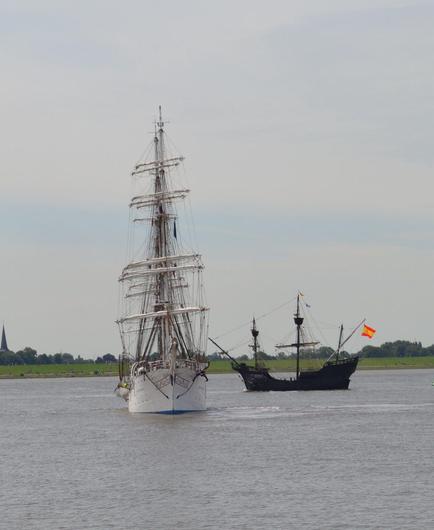 Weißes Segelschiff von vorn, daneben Nachbau eines Segelschiffs aus der Zeit um 1500 von der Seite. Beide Schiffe haben ihre Segel nicht gesetzt. 