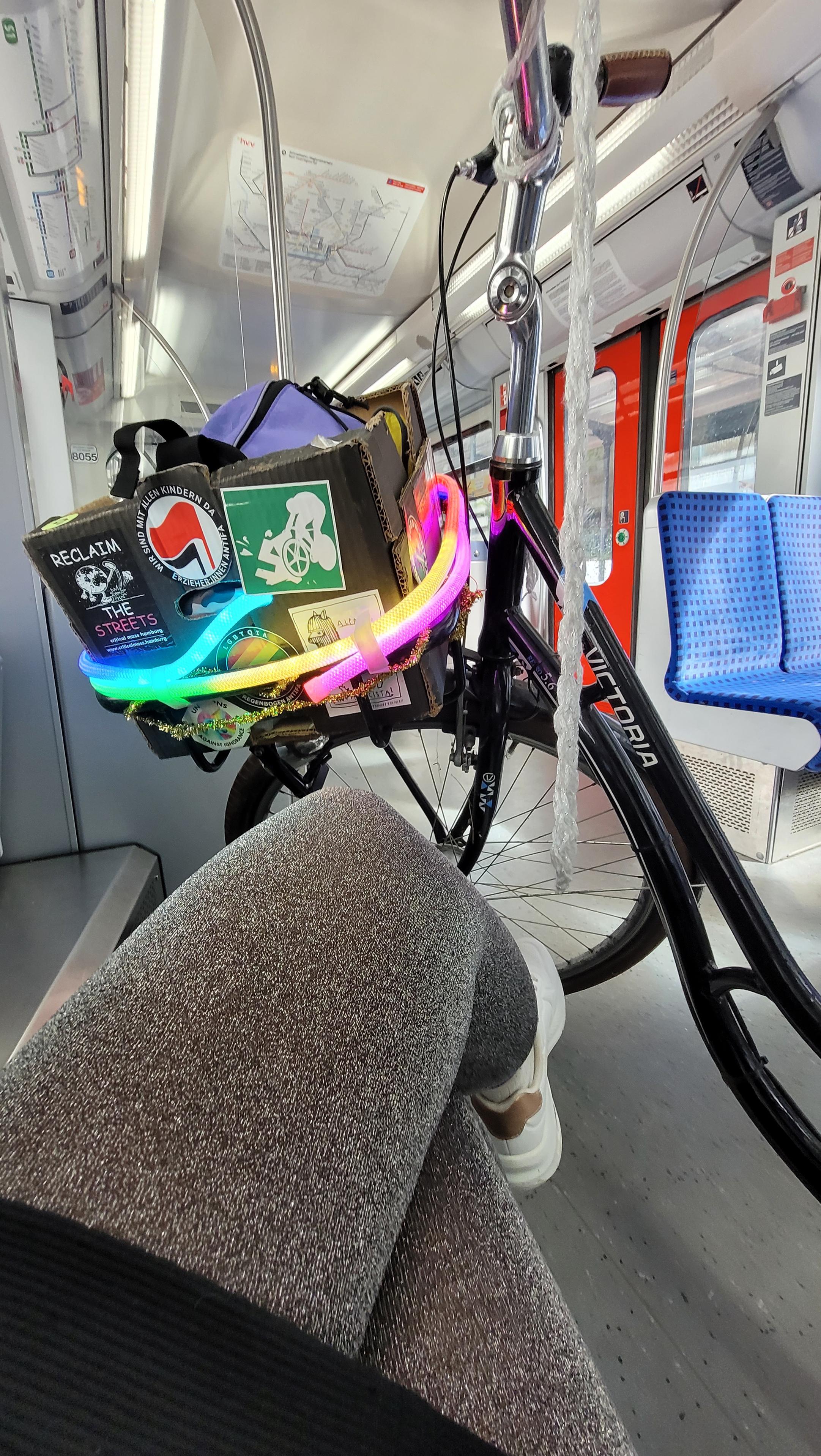 Hochkant in Farbe. Blick von der fotografierenden Person aus auf sitzende Beine in Glitzerstrumpfhosen und den vorderen Teil eines schwarzen Hollandrades. Vorne ist ein schwarzer Pappkarton als Korb angebracht, darauf verschiedene antifaschistische Sticker. Um den Korb leuchtet ein Lichterschlauch in Regenbogenfarben. Das Rad steht in einer s Bahn. Hochkant in Farbe. Blick von der fotografierenden Person aus auf sitzende Beine in Glitzerstrumpfhosen und den vorderen Teil eines schwarzen Hollandrades. Vorne ist ein schwarzer Pappkarton als Korb angebracht, darauf verschiedene antifaschistische Sticker. Um den Korb leuchtet ein Lichterschlauch in Regenbogenfarben. Das Rad steht in einer s Bahn.