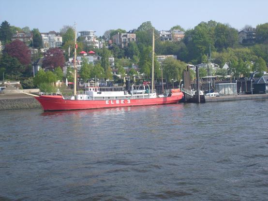 Schiff mit rotem Rumpf, beschriftet ELBE 3, an einem Ponton liegend. Im Hintergrund ein Hang mit Bäumen und Häusern.