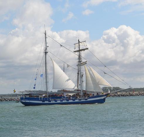 Ein zweimastiges Segelschiff, ein Teil der Segel ist gesetzt, im Hintergrund eine Mole.