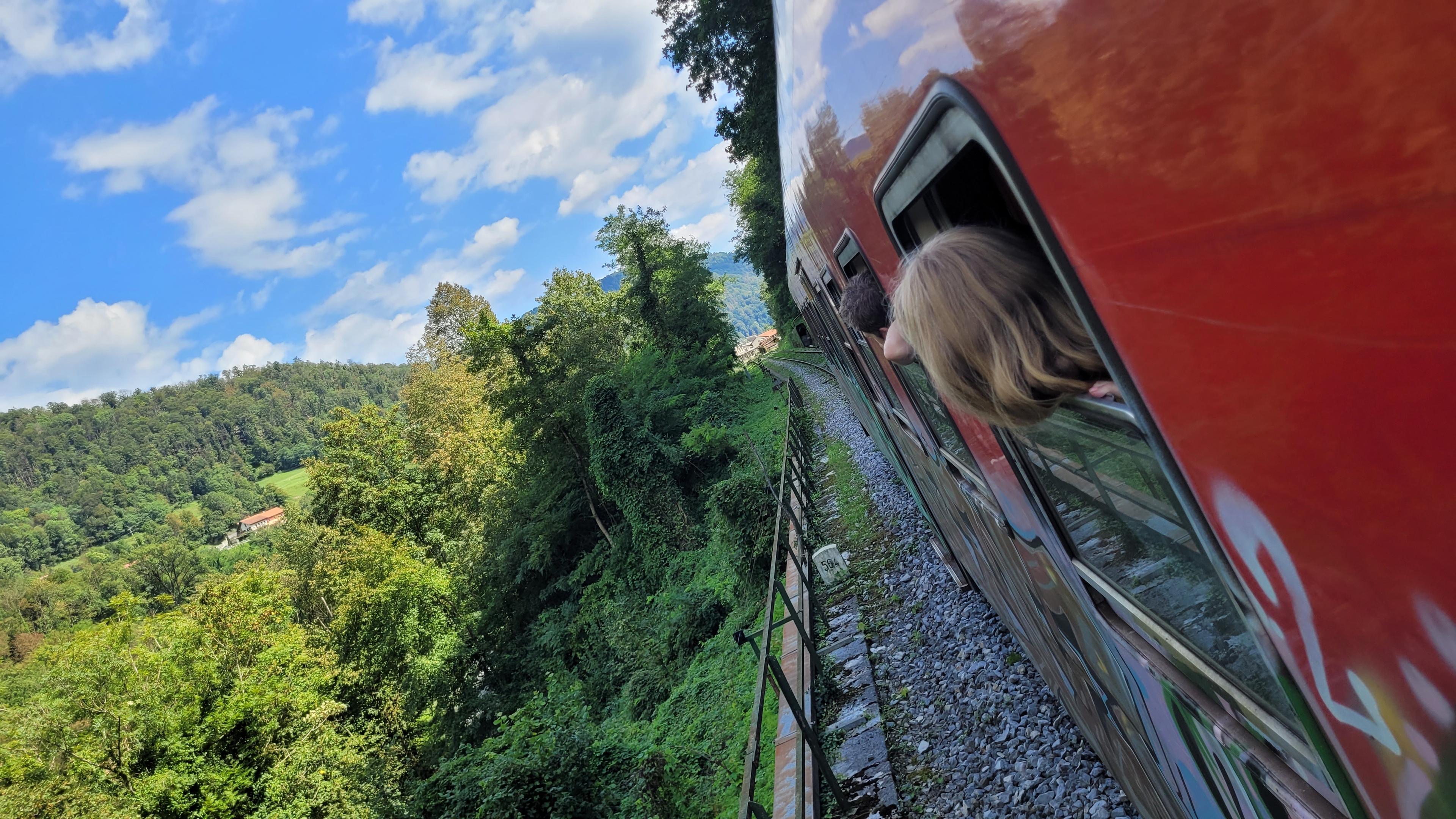 Querformat in Farbe. Schräg aufgenommen.  Blick einen roten Zug entlang. Aus den Fenstern schauen Menschen, man sieht ihren Hinterkopf. Am Zug entlang verläuft ein schmaler Streifen Schotter, abgegrenzt durch einen niedrigen Metallzaun. Am Ende der Schienen sind Häuser zu erkennen.
Links der Bahnlinie erstreckt sich ein Tal mit vielen Büschen und Bäumen. Alles ist sommergrün. Der Himmel ist blau mit Wölkchen. Das Rot des Zuges und das Grün des Tales grenzen sich kontrastreich voreinander ab.