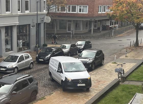 Ein Transporter steht mit eingeschalteter Warnblinkanlage quer über einen Parkplatz am Fahrbahnrand und auf dem angrenzenden Fußweg, den er zum überwiegenden Teil versperrt.