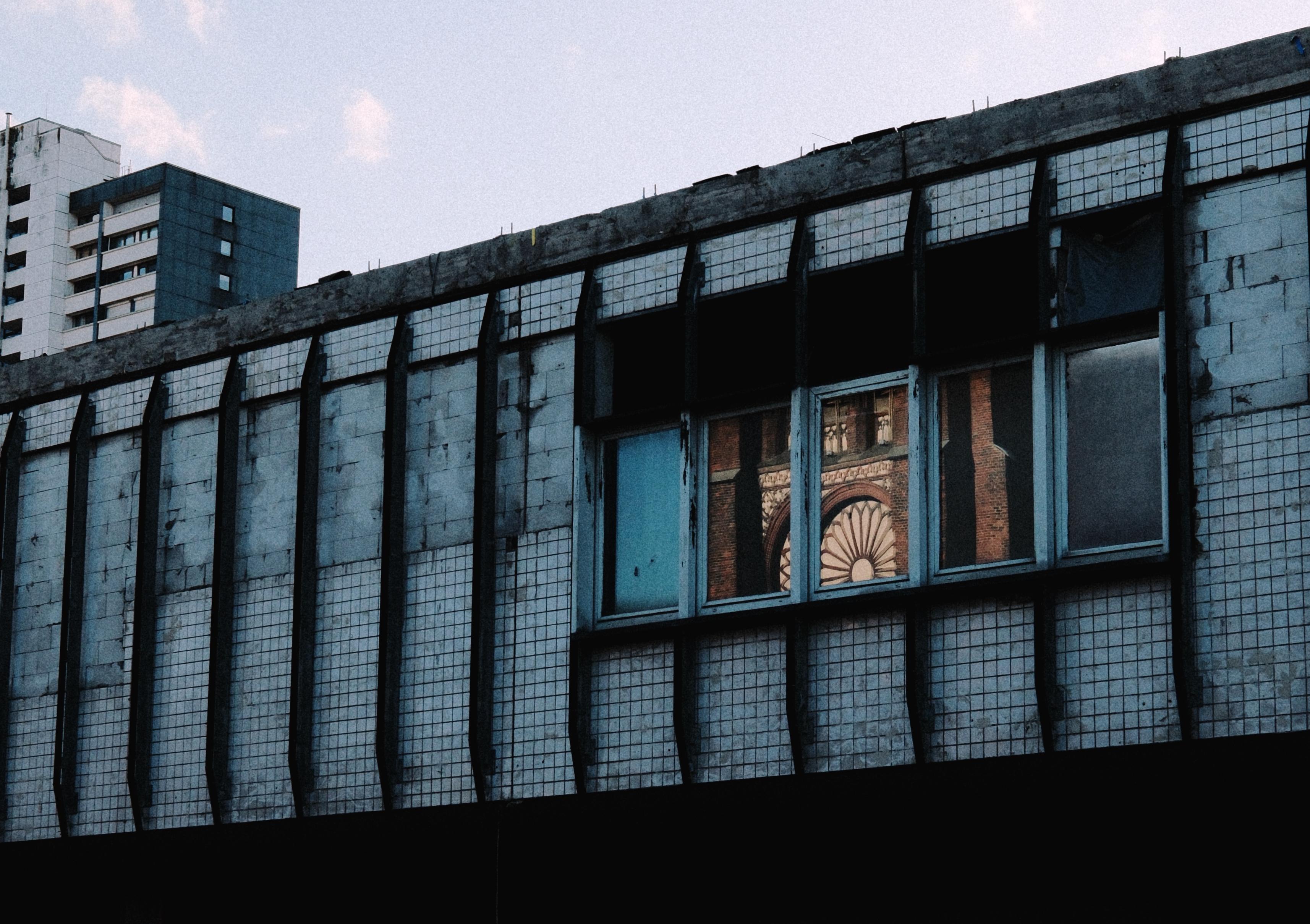 Ein städtisches Gebäude weist eine teilweise unterbrochene Fassade mit blauen Paneelen und gefliesten Abschnitten auf. Ein Fenster spiegelt ein detailliertes architektonisches Element eines benachbarten Gebäudes wider und zeigt ein geometrisches Muster. Der Himmel über dem Gebäude ist in sanften Farbtönen gefärbt.