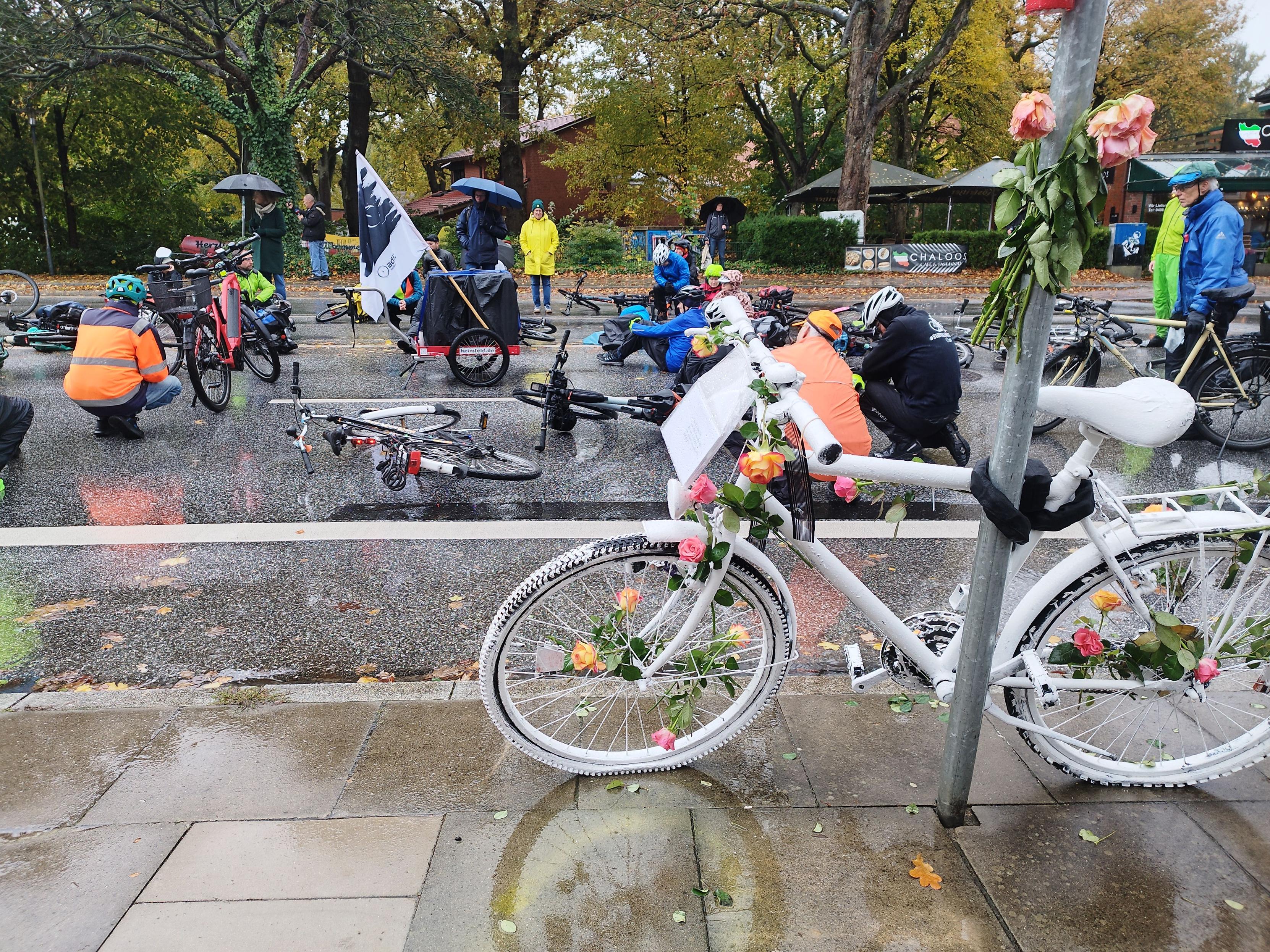 Menschen und ihre Räder auf einer Straße. Davor ein weiß angestrichenes Fahrrad, ein Ghostbike.
