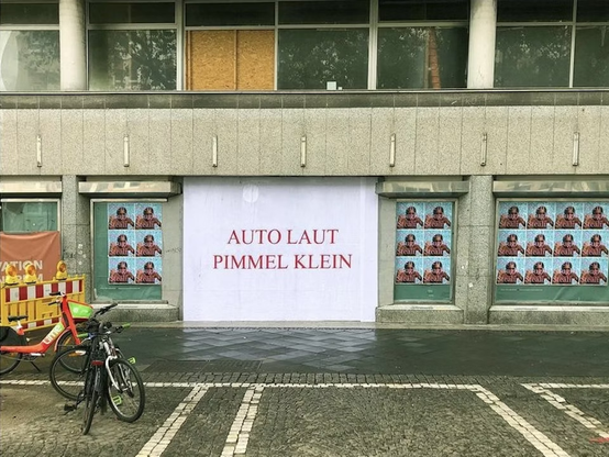 Foto einer Fassade mit einem großen weißen Werbeplakat "Auto laut, Pimmel klein"