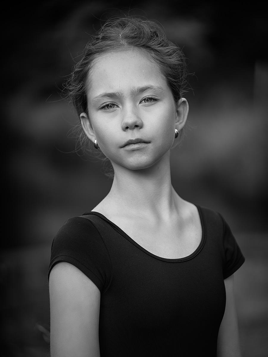A 12-year-old girl with her hair tied up is wearing a simple, short-sleeved black top. She stands against a blurred dark background and wears small, round earrings.