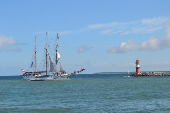 Dreimast-Segelschiff mit weißem Rumpf und teilweise gesetzten Segeln links im Bild, rechts eine Mole mit einem kleinen rot-weißen Leuchtturm.