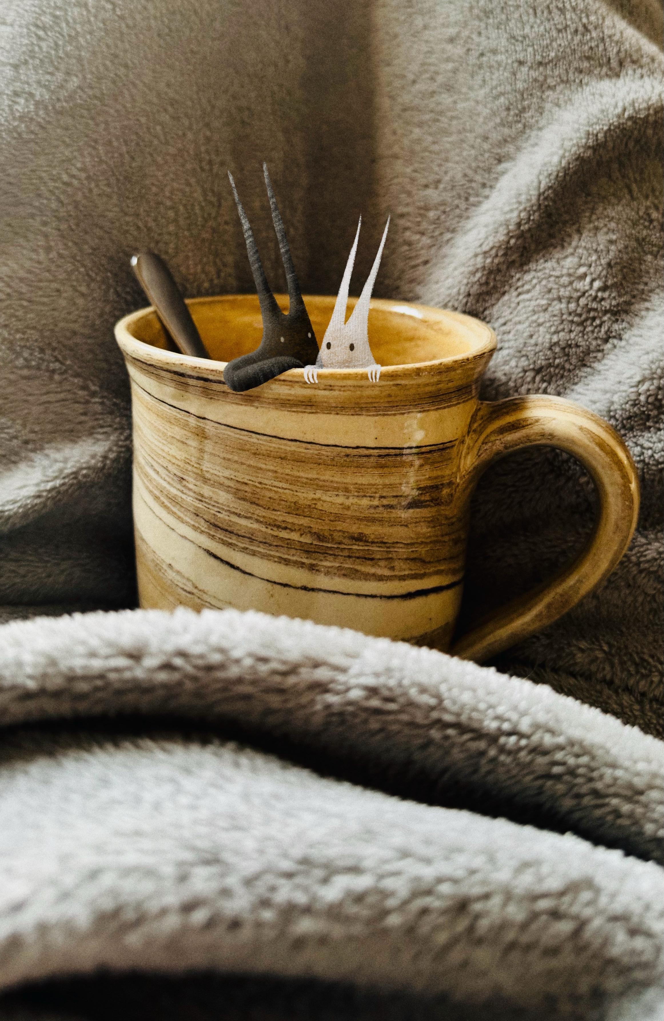 A cozy scene featuring a ceramic mug with two whimsical ghost-like figures inside, along with a spoon. The mug is placed on a soft, textured blanket, creating a warm and inviting atmosphere.