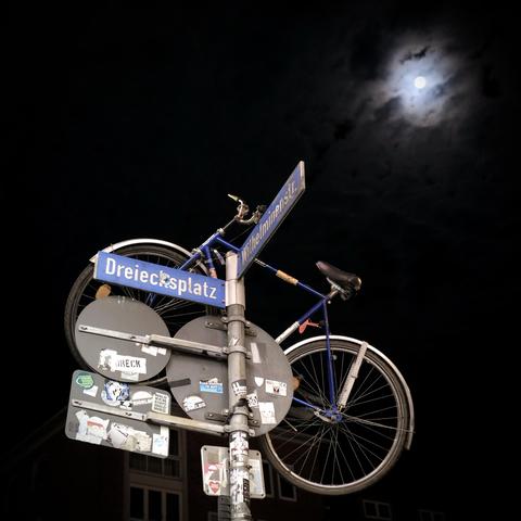 Schwarz dunkler Nachthimmel. Am Pfeiler von Straßenschildern hängt ein Fahrrad. Darüber scheint der Vollmond.