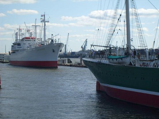 I'm Vordergrund der Heckteil eines grünen Segelschiffs, im Hintergrund ein weißes Frachtschiff schräg von vorne gesehen.