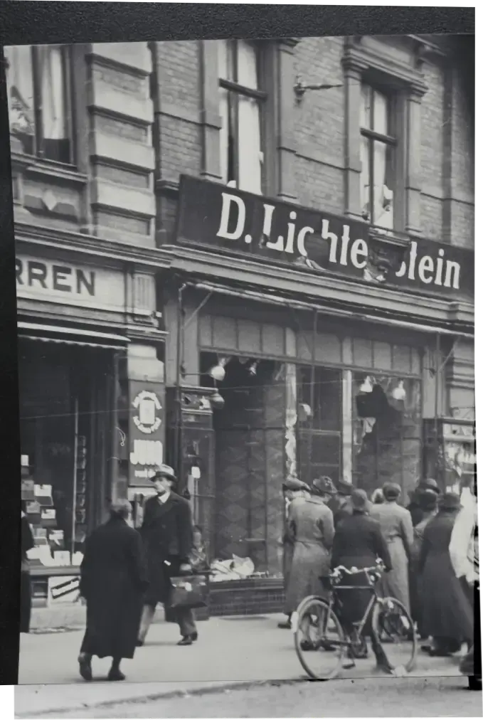 Schwarzweiß--Foto einer Ladenzeile. Zerbrochene Fenster am Laden von D. Lichtenstein. Menschen schauen in das Geschäft