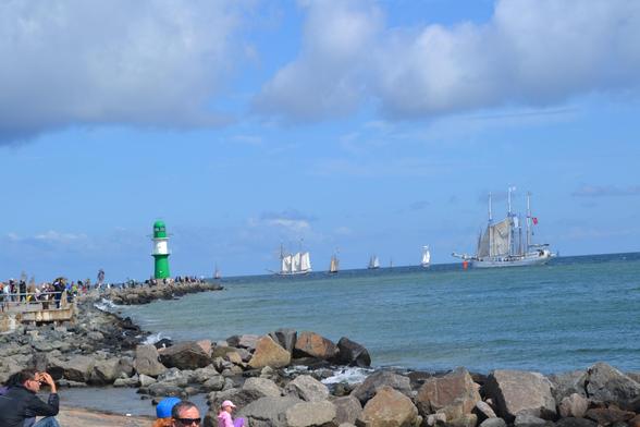Aus Felsbrocken bestehende Mole links im Bild, Zahlreiche Menschen auf der Mole. Am Ende der Mole ein kleiner grün-weiß gestreifter Leuchtturm. Rechts von der Mole das Meer mit einigen Segelschiffen.