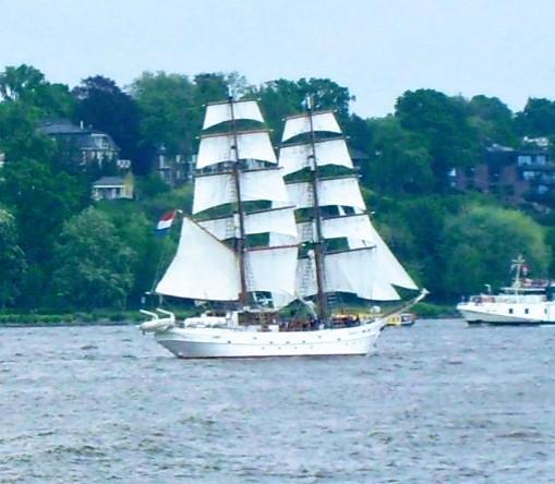 Weißes zweimastiges Segelschiff mit gesetzten Segeln, im Hintergrund ein baumbestandenen Ufer mit Villen.