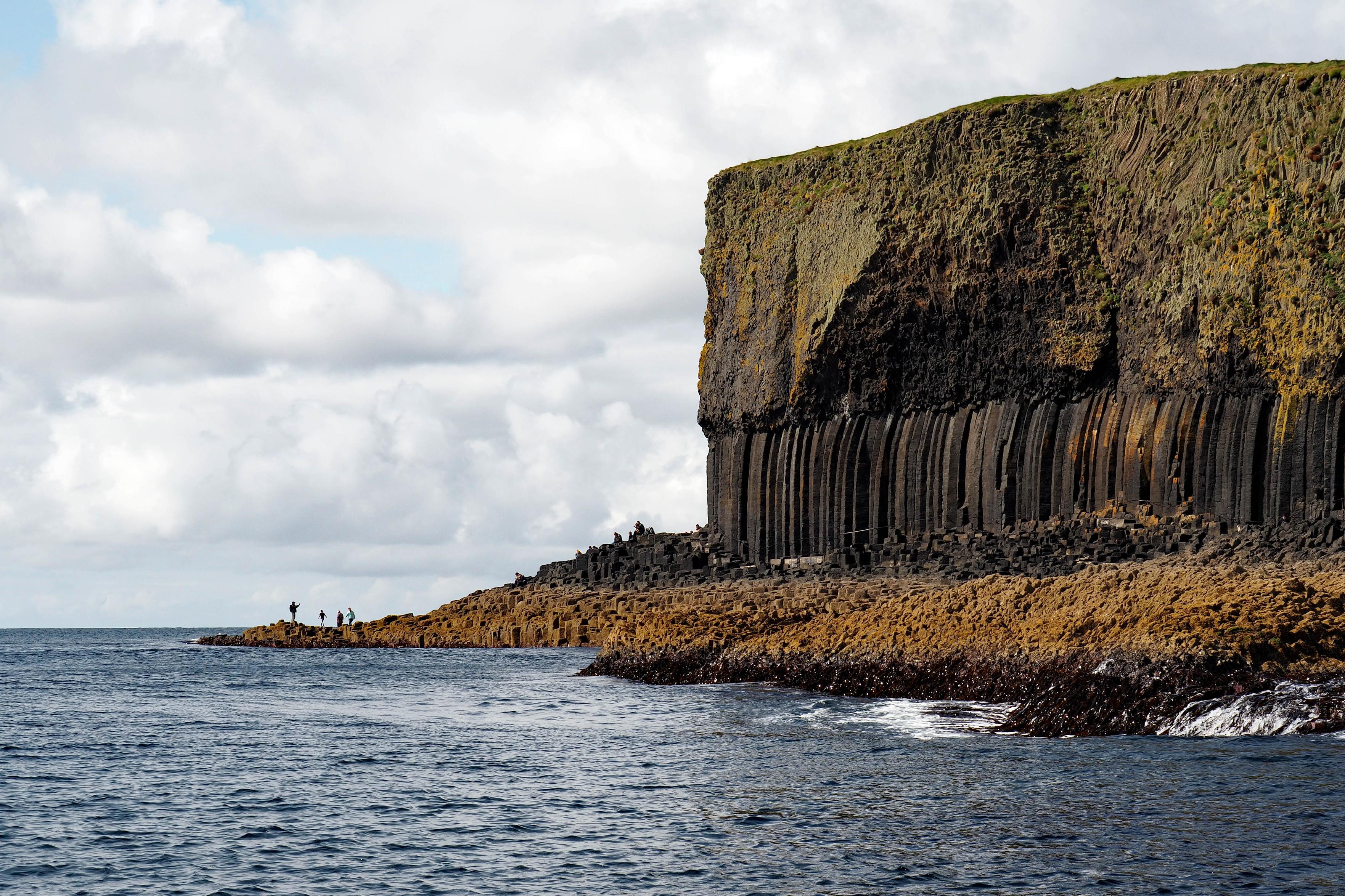 Eine Insel erhebt sich aus dem Meer, kleinere Wellen schlagen ans Ufer. Menschen stehen auf einer winzigen Landzunge und fotografieren die spektakuläre Gesteinsformation von Staffa. Es sind rechteckige Basaltsäulen aus Lavagestein, die eine doppelt so große Steinmasse tragen. Das Meer ist blau, der Himmel darüber mit vielen kleineren weißgrauen Wolken überzogen, gelborangefarbene Algen und ihre Ablagerungen färben die Uferzone. Das Gestein wechselt von grauschwarz zu grün-grau, mit Moosen und Flechten überzogen.
An island rises out of the sea, small waves lapping at the shore. People stand on a tiny headland and take photos of the spectacular rock formation of Staffa. These are rectangular basalt columns made of lava rock, supporting a stone mass twice their size. The sea is blue, the sky above is covered with many small white-gray clouds, and yellow-orange algae and their deposits color the shore. The rock changes from gray-black to green-gray, covered with moss and lichen.