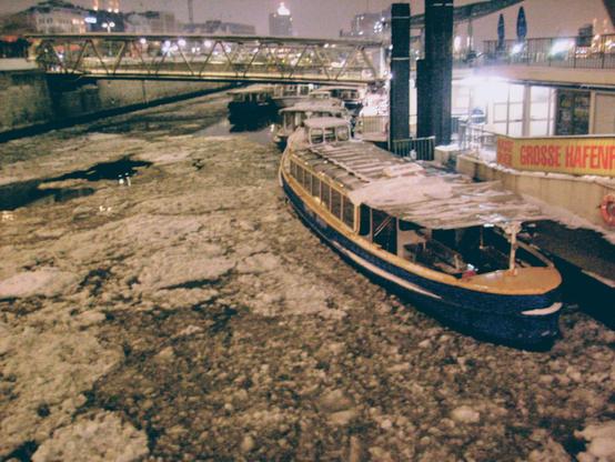 Die Innenseite der Sankt-Pauli-Landungsbrücken bei Nacht und strengem Frost. Rechts im Vordergrund eine Barkasse. Auf dem Wasser schwimmen Eisschollen.