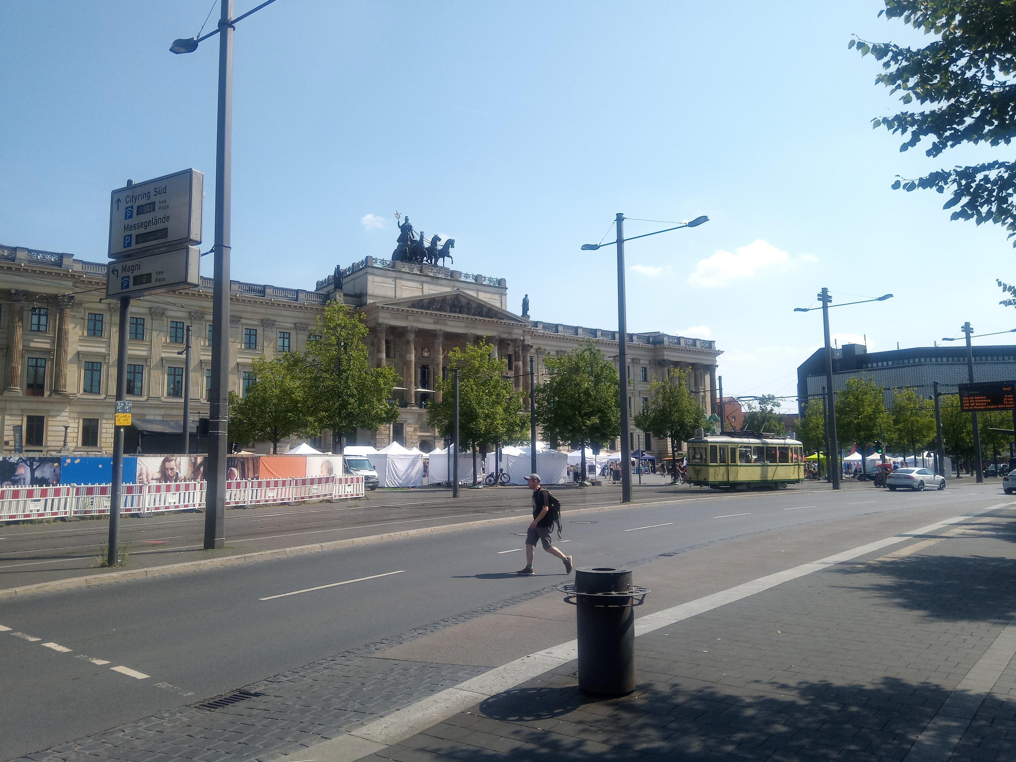 Eine zweispurige Einbahnstraße wird von einem Passanten an einer nicht dafür vorgesehenen Stelle überquert. Dann folgt eine Straßenbahnstrecke mit historischer Straßenbahn. Darauf die Gegenfahrbahn (kaum sichtbar) und dahinter viele Zelte. Dahinter eine Schlossfassade mit Quadriga (dahinter Einkaufszentrum) und rechts ein siebziger Jahre Kaufhaus (leer stehend). 