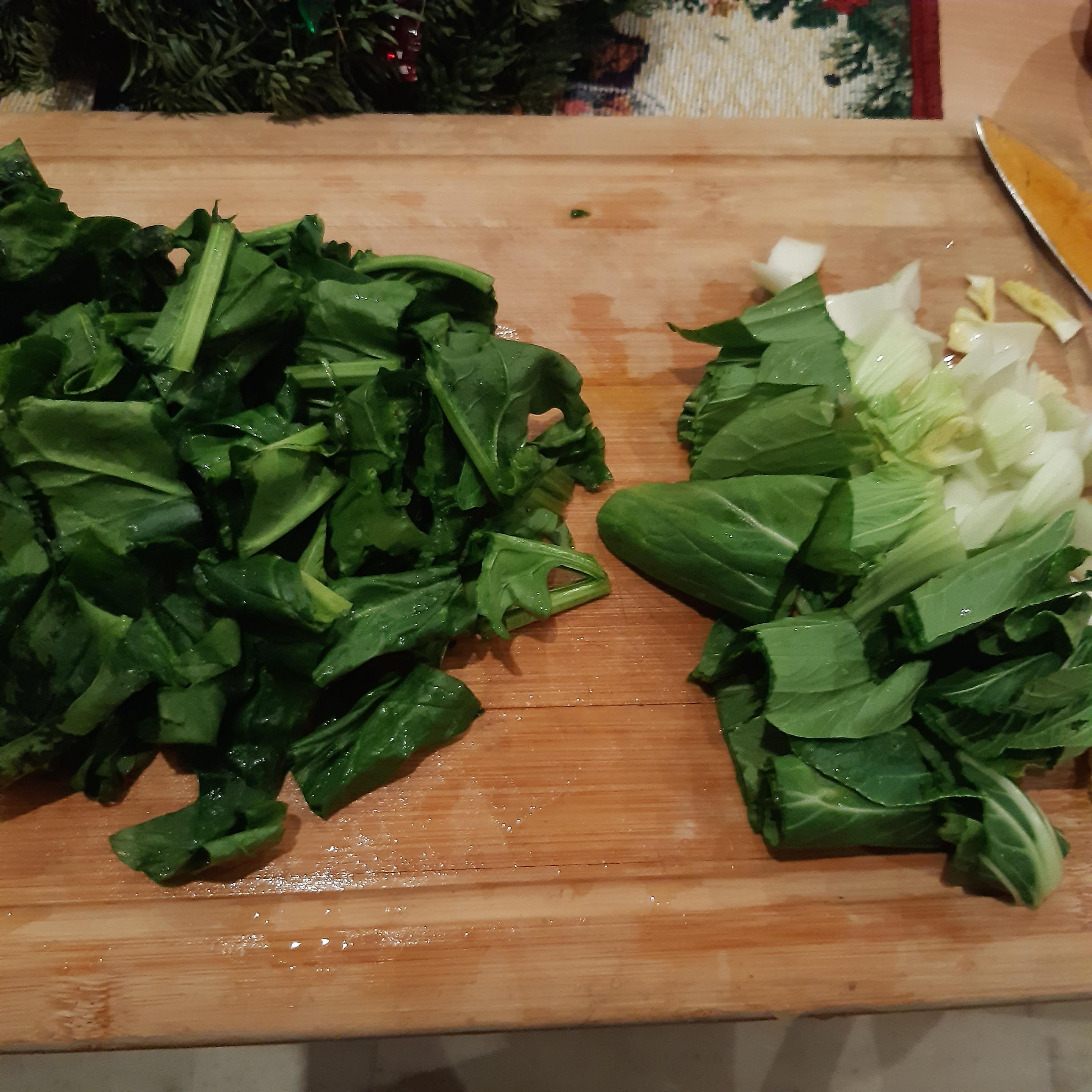Auf einem hölzernen großen Küchenbrett liegt auf der linken Seite kleingeschnitterner Spinat und auf der rechten Seite kleingeschnittener Pak Choi.