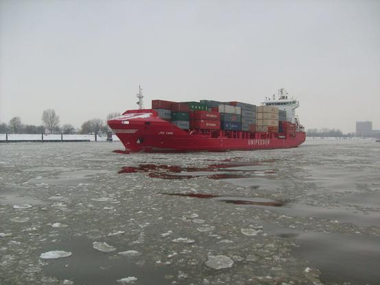 Containerschiff mit rotem Rumpf, beladen mit Containern, hoch an Deck gestapelt. Das Wasser mit kleinen Eisschollen bedeckt.