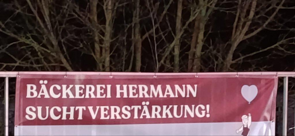 An einen Geländer ein Transparent mit den Worten in Großbuchstaben "Bäckerei Hermann sucht verstärkung"
Das leer Zeichen zwischen den letzten beiden Worten ist aber nicht wirklich zu erkennen.