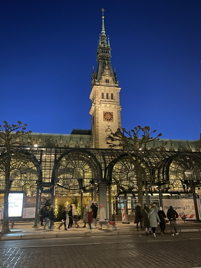 Im Vordergrund Menschen unter bogenförmigen Glasdächern. Dahinter/darüber das Rathaus aus Sandstein, angestrahlt, vor noch nicht ganz dunklem Himmel.