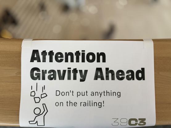 A printed sign on a wooden railing reads "Attention Gravity Ahead" with a warning not to put anything on the railing. An illustration of an object falling is included. The bottom right corner features "39c3." The background is blurred showing a busy crowd. 