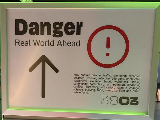 A warning sign labeled "Danger: Real World Ahead" with an upward arrow and an exclamation mark. It lists various potential experiences and issues associated with the real world, including people, traffic, weather, friendship, and more.