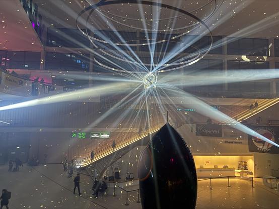 An indoor space featuring modern architecture with a large circular light fixture. Bright beams of light radiate from the center, creating a dramatic effect. The area is bustling with people, and there are signs for directions and information in the background.